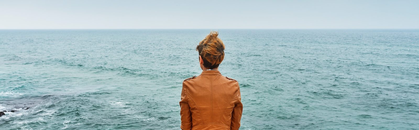 woman in brown jacket sitting on brown rock near sea during daytime