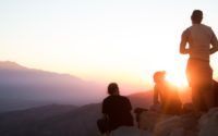 two men sitting and one man standing near cliff taken during golden hour